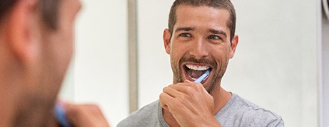 A happy man brushing his teeth in front of a bathroom mirror