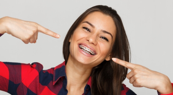 Young woman with traditional braces pointing to her smile