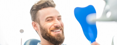 Bearded man in dental chair smiling while holding a mirror