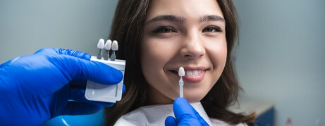 Young woman in dental chair with dentist holding tooth color chart to her smile